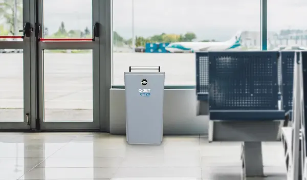 Grauer Abfalleimer mit Aufschrift 'Q-JET CT20' steht vor Glastür in Flughafenwartebereich mit blauen Sitzbänken und Flugzeug im Hintergrund.