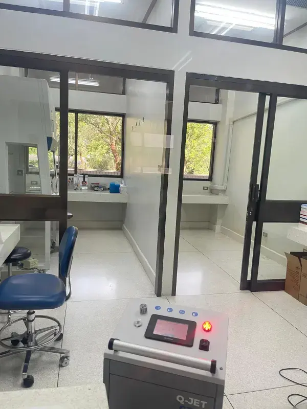 Laborraum mit weißen Wänden, mehreren Fenstern, einem blauen Drehstuhl und einem Q-JET-Gerät mit beleuchtetem roten Knopf.