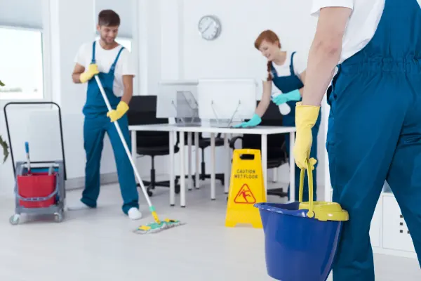 Drei Reinigungskräfte in blauen Overalls und gelben Handschuhen reinigen ein Büro mit Schreibtischen und Computern, Warnschild steht auf dem Boden.
