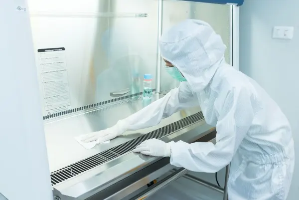 Person in weißem Schutzanzug, Handschuhen und Mundschutz reinigt sterile Arbeitsfläche im Labor unter Abzugshaube.
