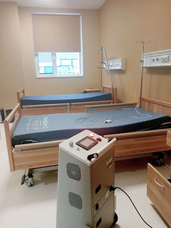 Zwei Krankenhausbetten mit blauen Matratzen und zwei Infusionsständern in einem hellen Patientenzimmer mit geschlossenem Fenster und einem medizinischen Gerät im Vordergrund.