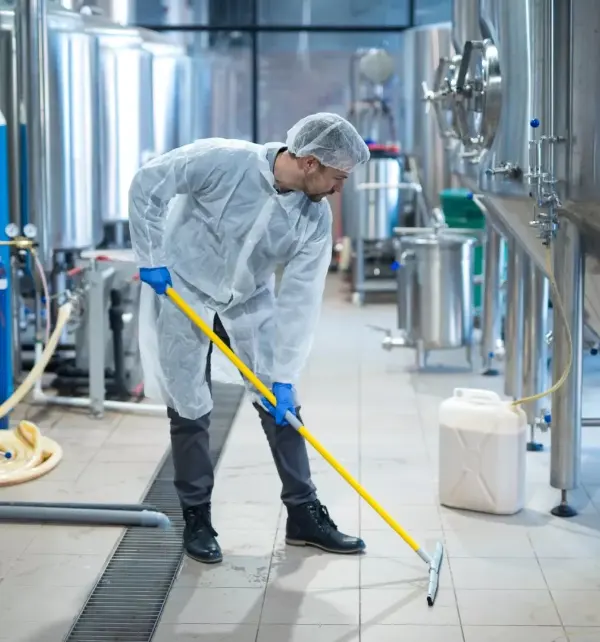 Person in Schutzkleidung und Haarnetz reinigt Boden mit gelbem Wischmopp in Lebensmittelproduktionsanlage.
