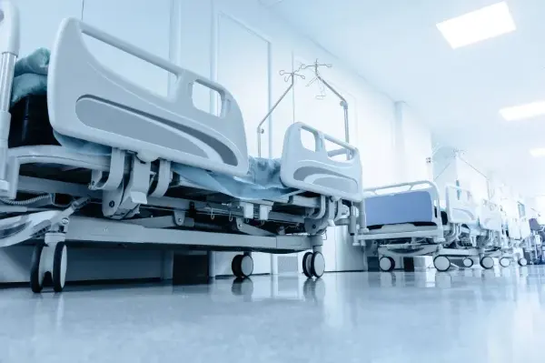 Zwei leere Krankenhausbetten mit weißen Metallrahmen und Rollen in einem hellen Krankenzimmer mit Infusionsständern.