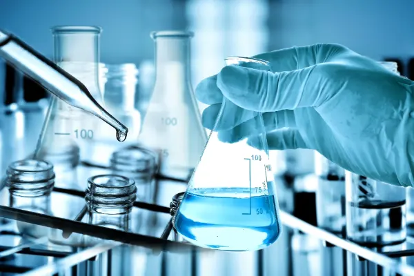 Hand mit Schutzhandschuh hält einen Erlenmeyerkolben mit blauer Flüssigkeit, daneben Tropfpipette mit Tropfen über Glasgefäß, Laborutensilien im Hintergrund.