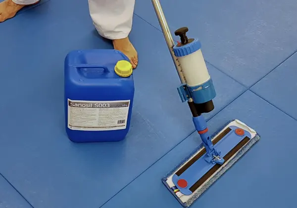 Blauer Kanister mit gelbem Verschluss und Aufschrift 'Sanosil S003' neben einem Wischmopp auf blauem Boden, daneben eine Person mit weißen Hosen und nackten Füßen.