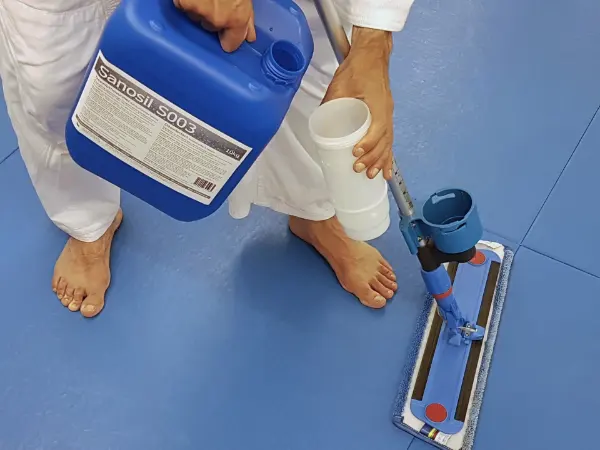 Barfüßige Person in weißem Anzug gießt Flüssigkeit aus blauem Kanister mit Aufschrift 'Sanosil S003' in weißen Messbecher über blauem Boden mit Wischmopp.