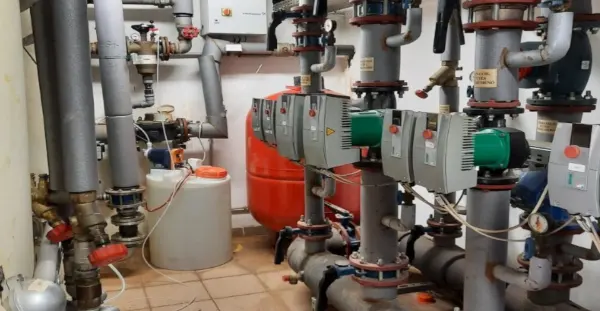 Rohrleitungen und Pumpen in einem Technikraum, darunter mehrere vertikale Rohre mit Ventilen und Messgeräten, ein roter Druckbehälter und ein weißer Kunststoffbehälter.