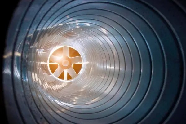 Blick in ein rundes Lüftungsrohr mit spiralförmiger Innenstruktur und einem Ventilator am Ende, durch den Sonnenlicht scheint.