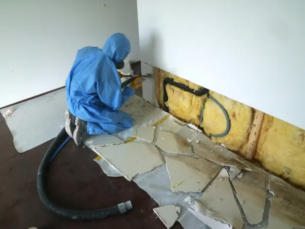 Person in blauem Schutzanzug kniet vor einer Wand mit freigelegter Dämmung und entfernt beschädigte Wandverkleidung.