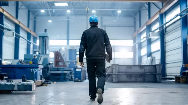 Person mit blauem Schutzhelm und dunkler Arbeitskleidung geht in einer großen Industriehalle weg, Blick von hinten.