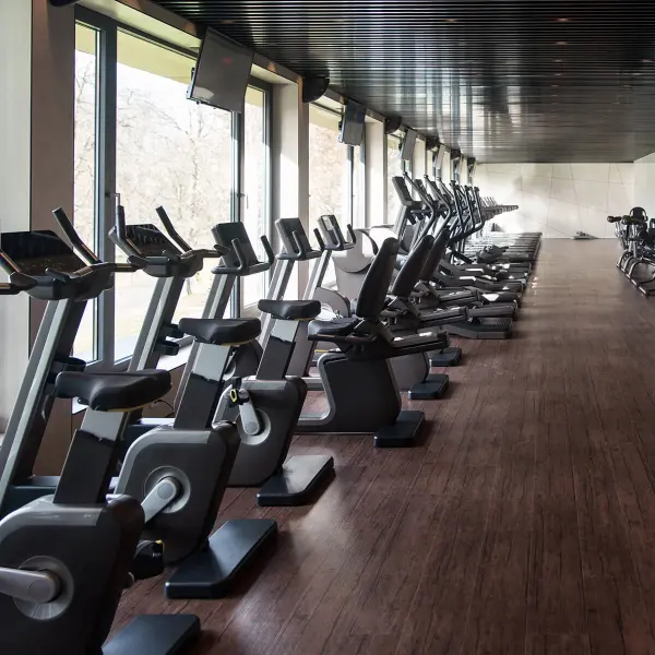 Reihe von stationären Fahrrädern mit Monitoren in einem Fitnessraum mit großen Fenstern und Holzboden.